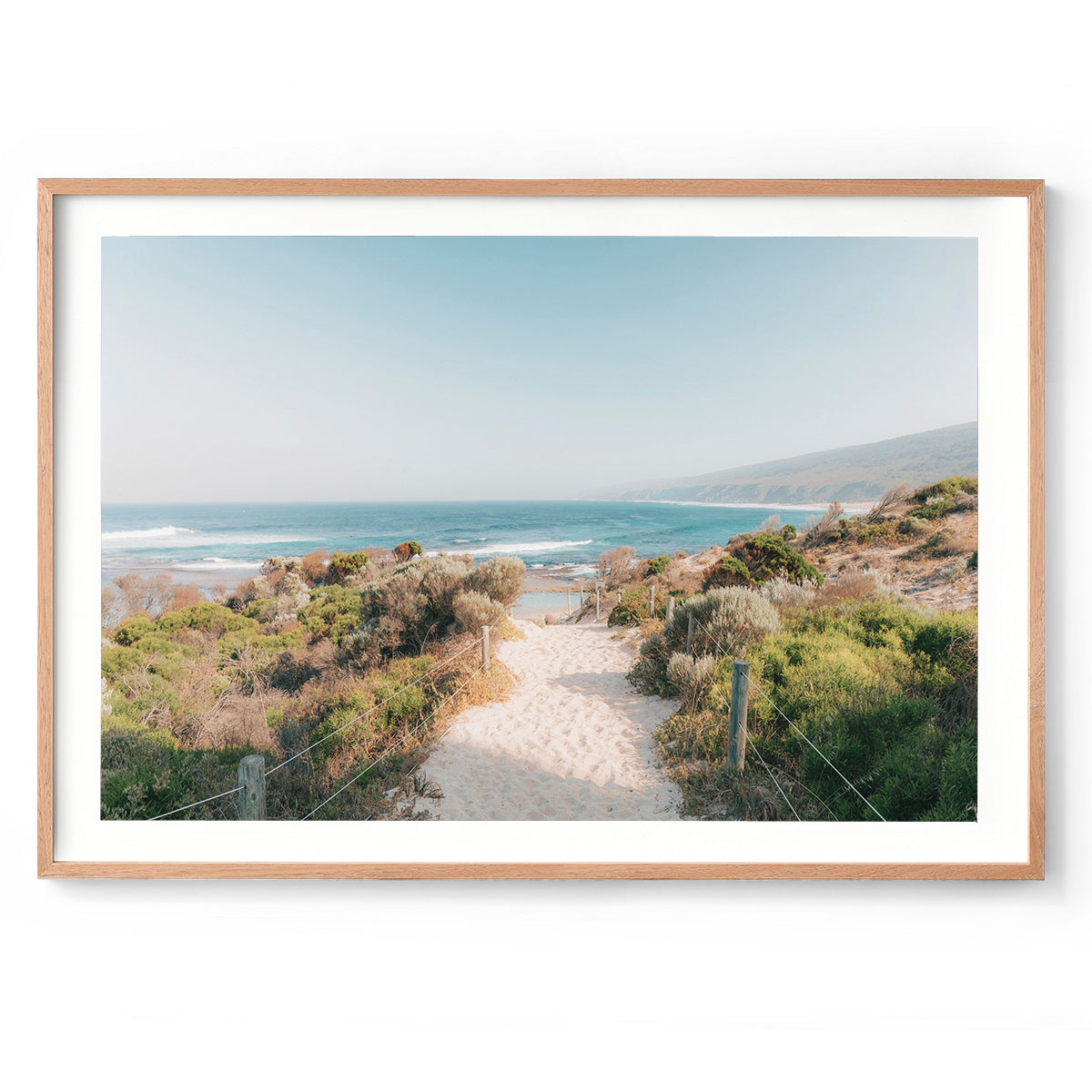 Framed photo of the sandy beach path leading down towards Yallingup Beach in Western Australia. The bush frames the path with the blue ocean and cliffs at the end. Available framed in black, white or oak.