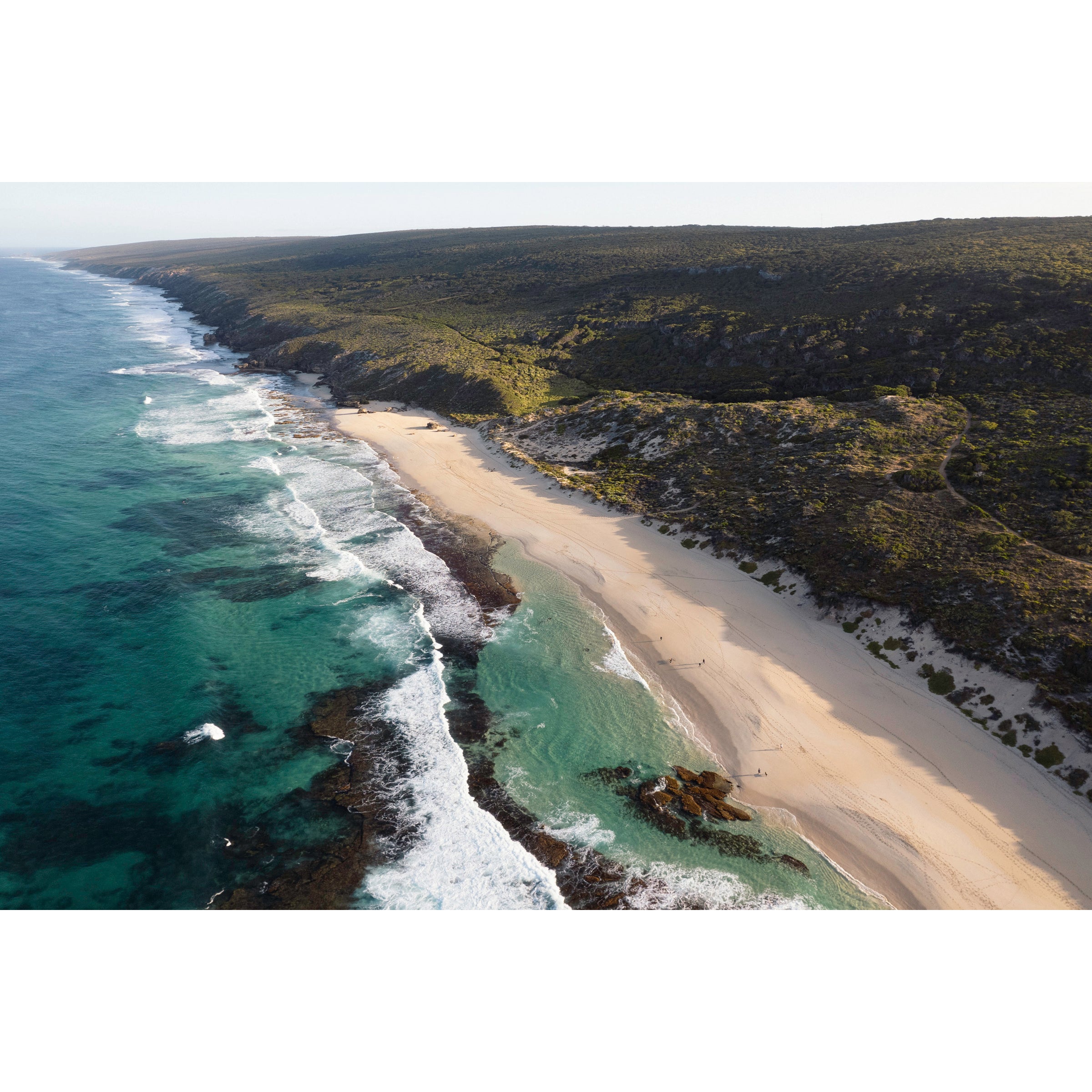 Horizontal drone photo of the far end of Yallingup Beach with the waves rolling in to the white sand and the bushland stretching into the distance. Original fine art photography of Western Australia, available as prints and framed wall art. Perfect for a coastal home or beach house.