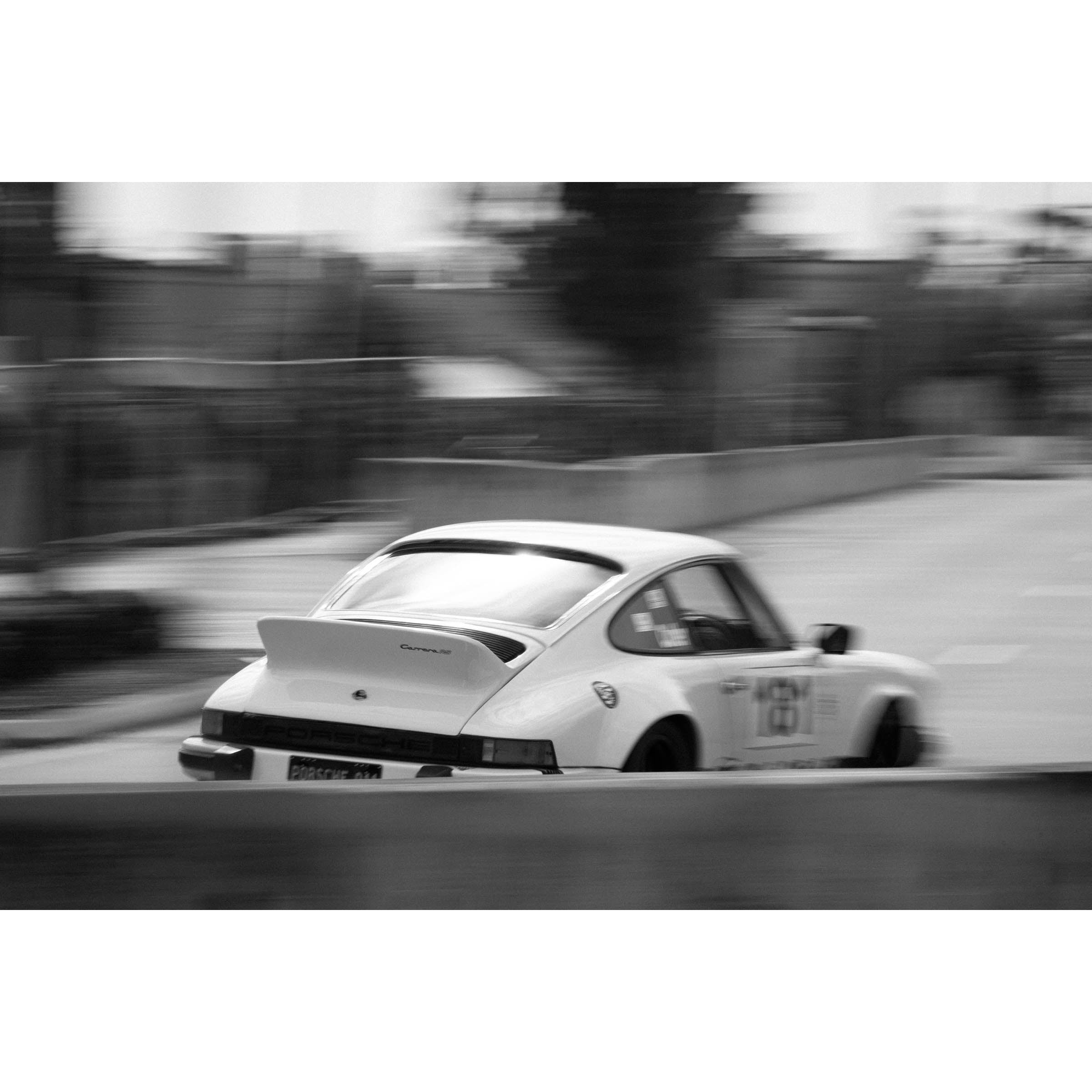 Horizontal black and white photo of a white Porsche 911 racing in the streets, the background is blurred as it goes around a corner. Fine art photography prints, available framed in black, white or oak.