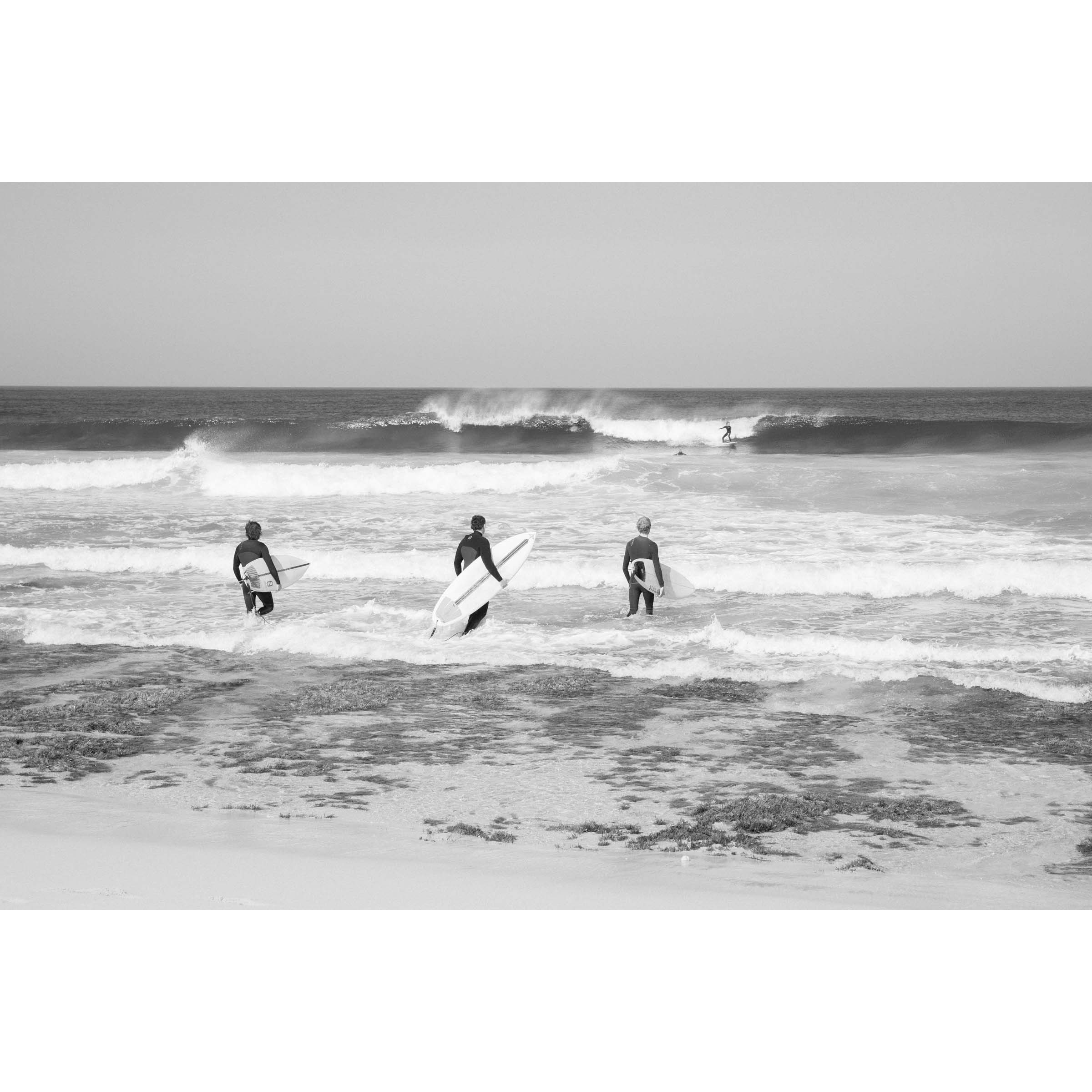 Horizontal black and white photo of three surfers walking into the ocean at Rabbits Break, Yallingup Beach, Western Australia. They have their surfboards under their arms and a wave rolls in with a surfer on it. Fine art photography prints, available framed in black, white or oak.