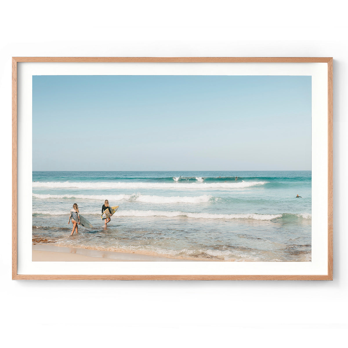 Framed horizontal photo of two female surfers walking into the ocean at Yallingup Beach in Western Australia holding their surfboards under their arms. Waves are rolling in with surfers dotted throughout the soft blue water. Available framed in oak, black or white.
