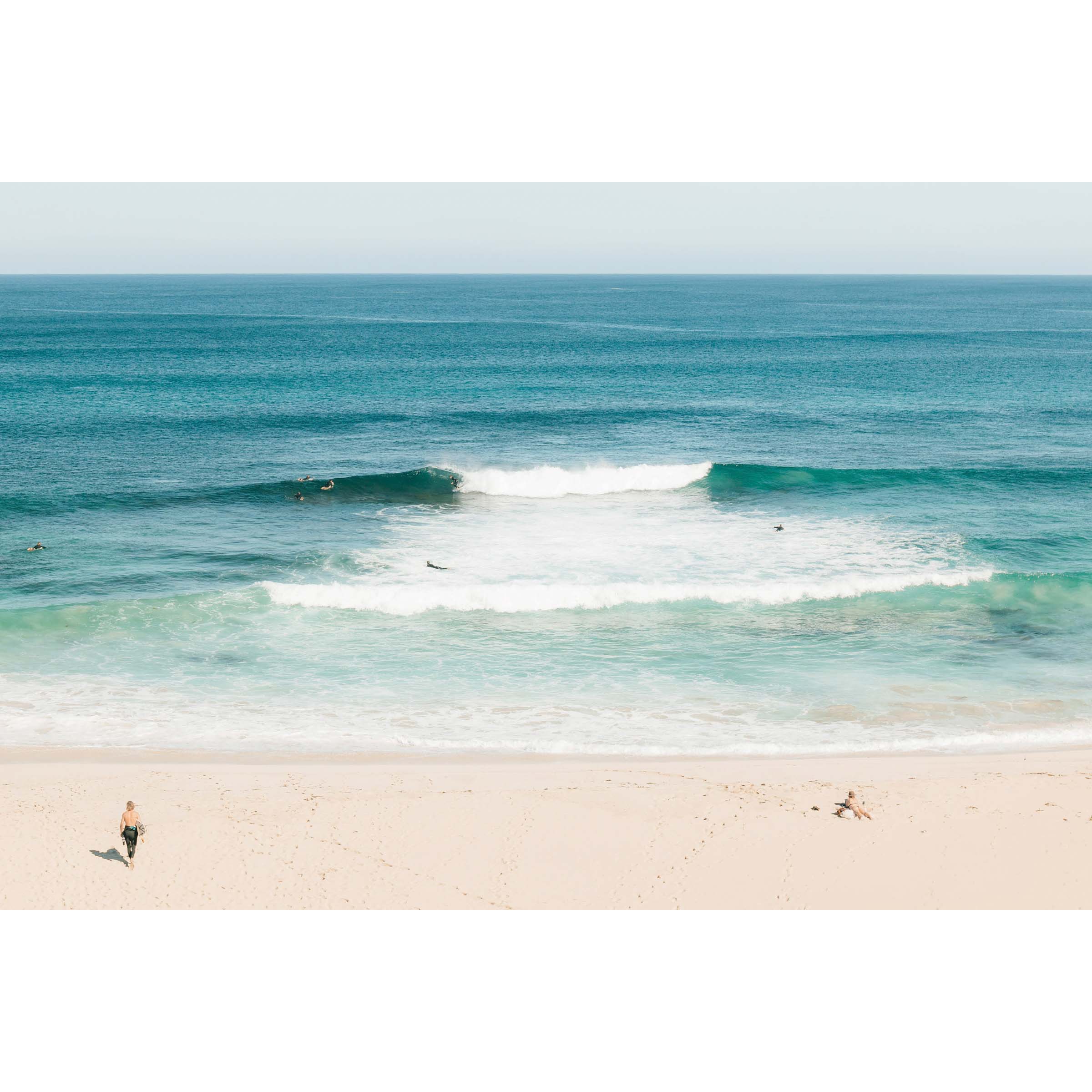 Framed photo of Injidup Beach in Yallingup in Western Australia. There are waves rolling in with surfers dotted through the ocean and a surfer walks across the sand to the water with a surfboard under his arm as a girl sunbathes. Available framed in black, white or oak.