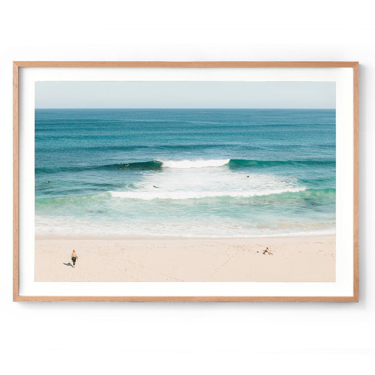 Framed photo of Injidup Beach in Yallingup in Western Australia. There are waves rolling in with surfers dotted through the ocean and a surfer walks across the sand to the water with a surfboard under his arm as a girl sunbathes. Available framed in black, white or oak.