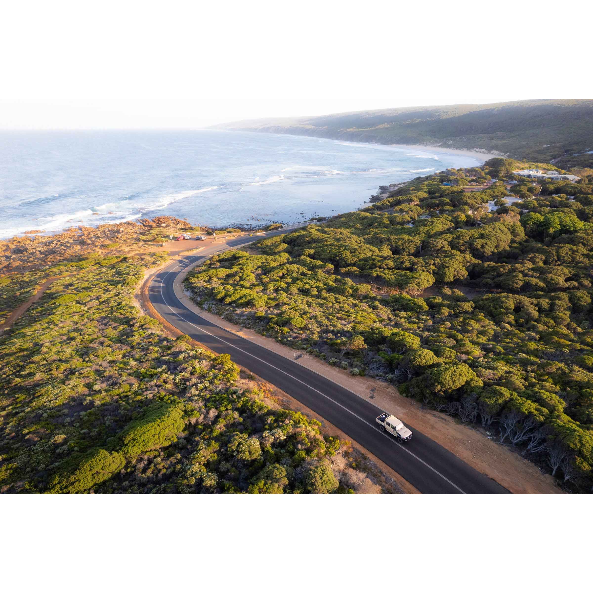 Horizontal photo of the road down to Yallingup beach in Western Australia. Taken from above at sunrise so it has a golden glow, with a car driving up the hill and the ocean in the background. Perfect for the coastal home or beach house. Available as fine art photography prints and framed wall art.