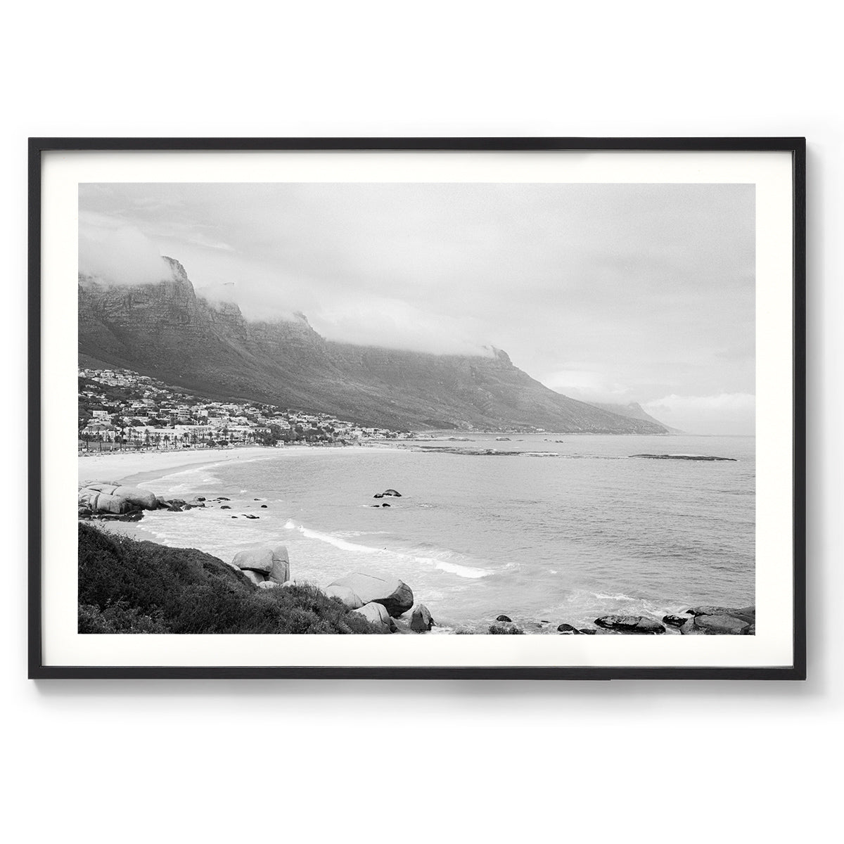 Framed black and white photo of Camps Bay in Cape Town, South Africa. Shot on film on a moody day, the clouds are covering the tops of the mountains and gentle waves roll in as the city curves around the beach. Available framed in black, white or oak.