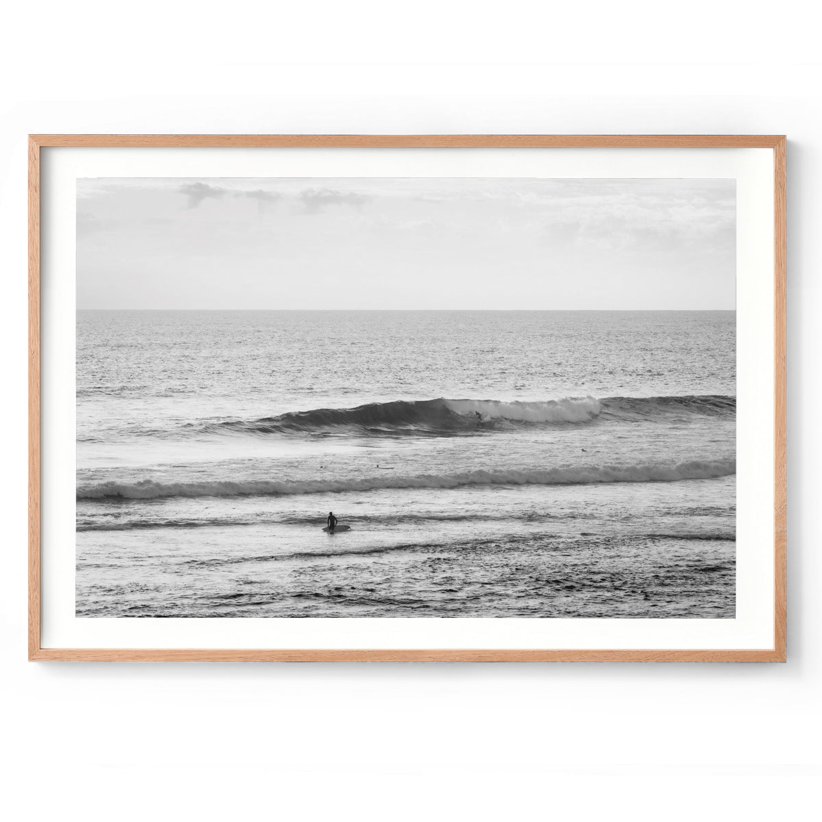 Black and white minimalist photo of a person walking out of the ocean holding a surfboard at Yallingup Beach in Western Australia. A small surfer is on a wave in the background. Fine art photography prints, available framed in black, white or oak.