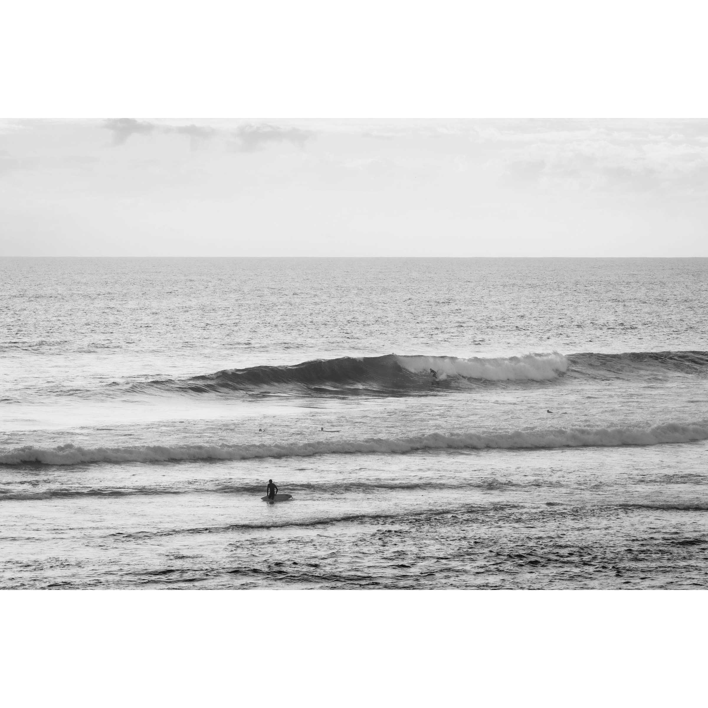 Black and white minimalist photo of a person walking out of the ocean holding a surfboard at Yallingup Beach in Western Australia. A small surfer is on a wave in the background. Fine art photography prints, available framed in black, white or oak.