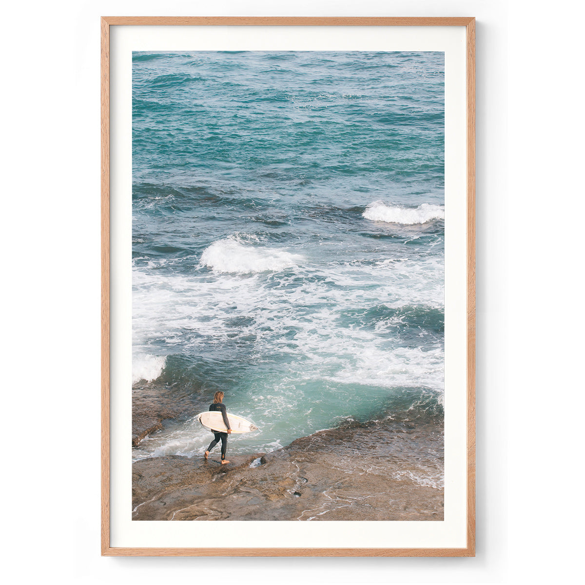 Person holding a surfboard on a rocky shore with ocean waves in the background. Shot at Surfer's Point in Margaret River, Western Australia. Available framed in black, white or oak.