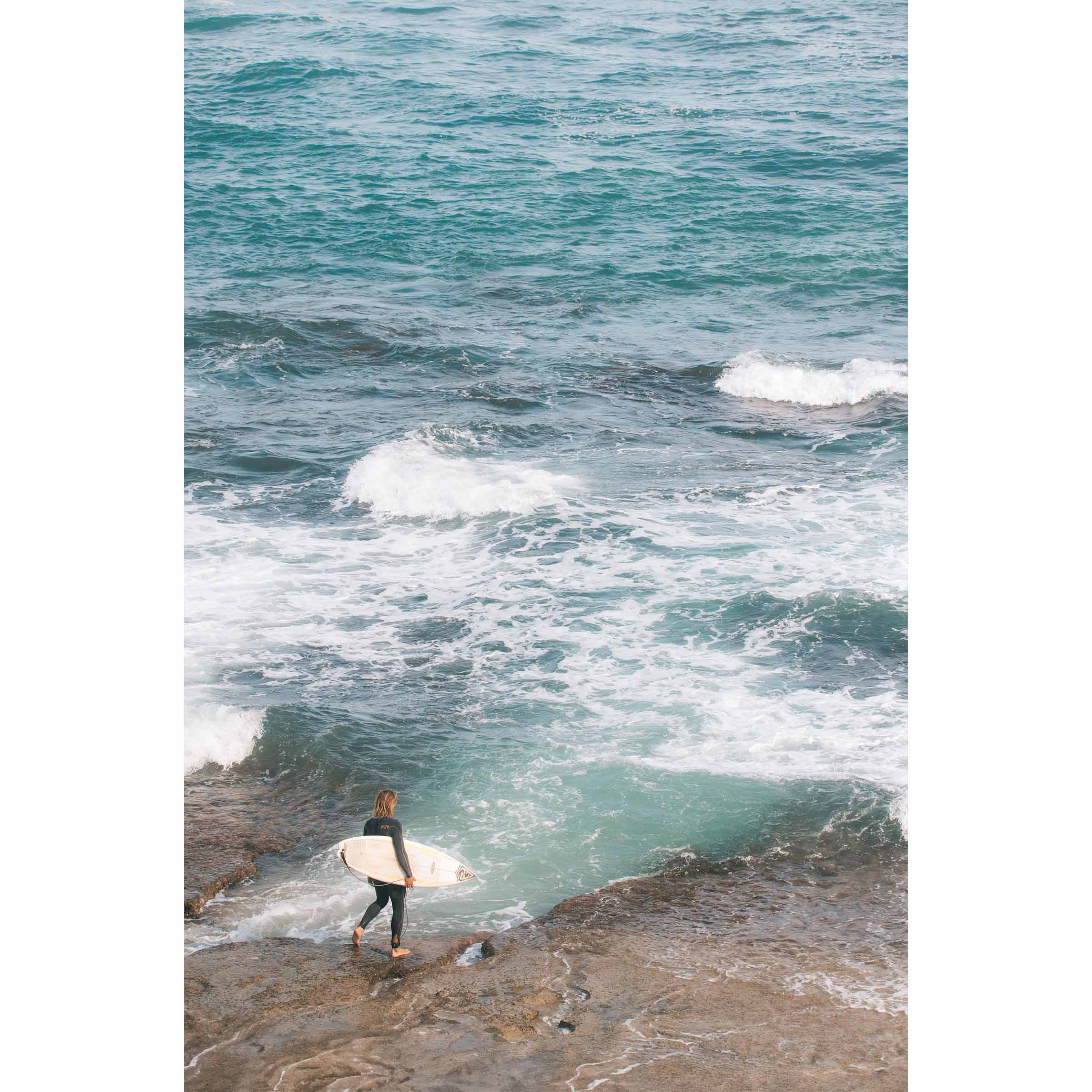 Person holding a surfboard on a rocky shore with ocean waves in the background. Shot at Surfer's Point in Margaret River, Western Australia. Available framed in black, white or oak.