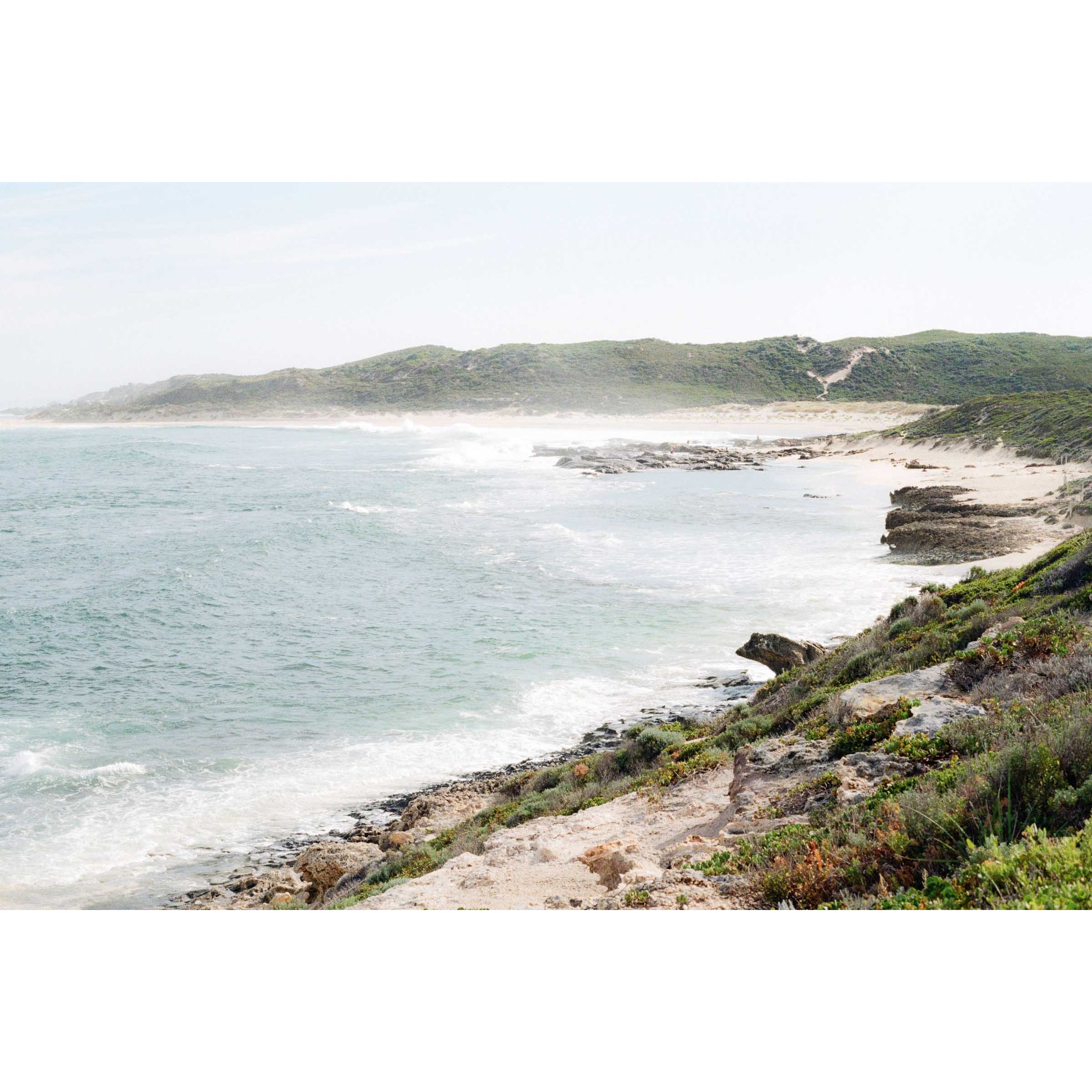 Photo of a coastal landscape at Surfer's Point in Margaret River, Western Australia. Soft ocean waves roll in with the bush and rocks curving around. The spray from the ocean makes it a bit dreamy, as well as soft colours as it's shot on film. Available framed in black, white or oak.