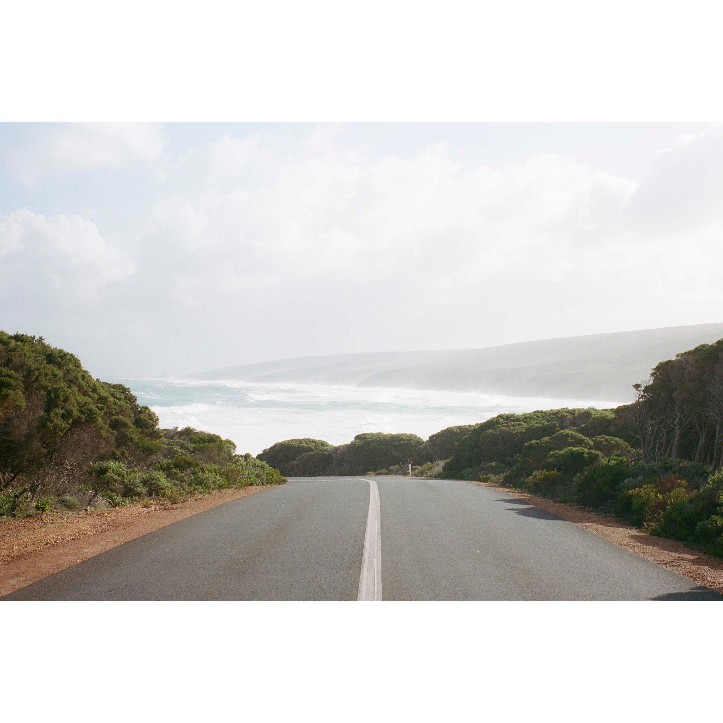 Horizontal photo of the road leading down to Smiths Beach in Yallingup, Western Australia. Shot on film it has muted colours with the bush either side of the road and the water and hills in the distance. Original Australian photography, available as fine art prints and framed wall art. Perfect for the coastal home or beach house.