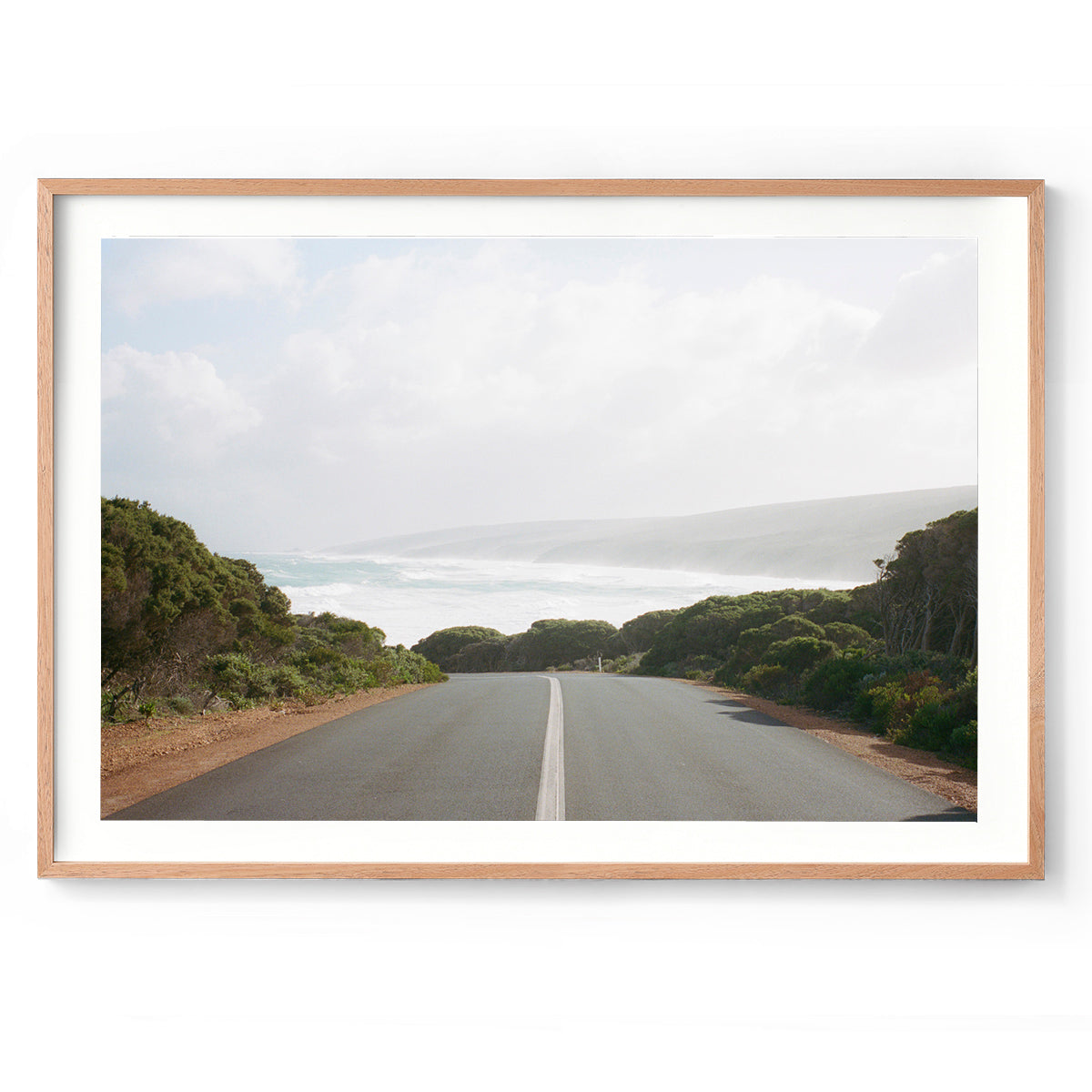 Horizontal photo of the road leading down to Smiths Beach in Yallingup, Western Australia. Shot on film it has muted colours with the bush either side of the road and the water and hills in the distance. Original Australian photography, available as fine art prints and framed wall art. Perfect for the coastal home or beach house.