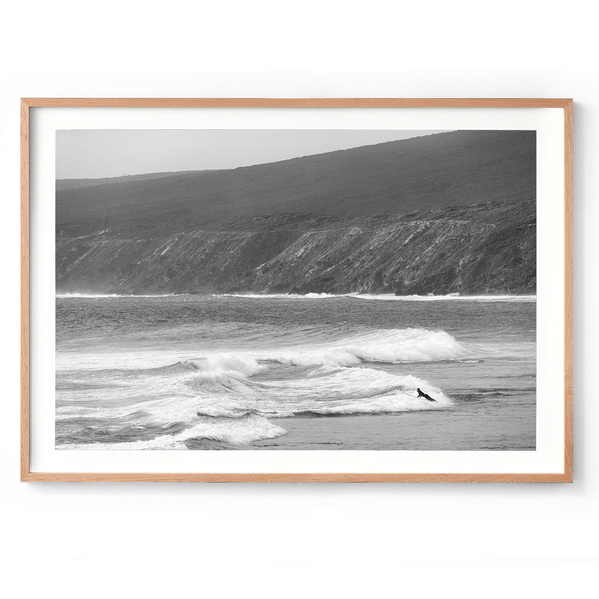 Horizontal black and white photo of a person mid air as he moves onto his surfboard in the ocean to paddle out at Yallingup Beach in Western Australia. The iconic cliffs of Rabbit Hill are in the background. It has a nostalgic feel. Fine art photography prints, available framed in black, white or oak.