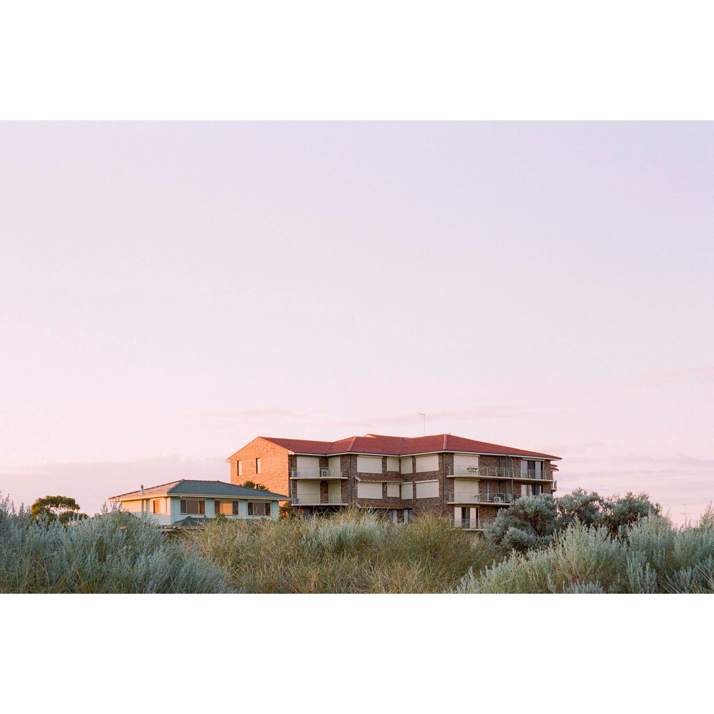 Horizontal photo of the top of a brick 80s block of units in Mandurah, Western Australia. Most of the photo is a soft, lavender sky with silver green bushes in the foreground. Original fine art photography, available as prints and framed wall art.