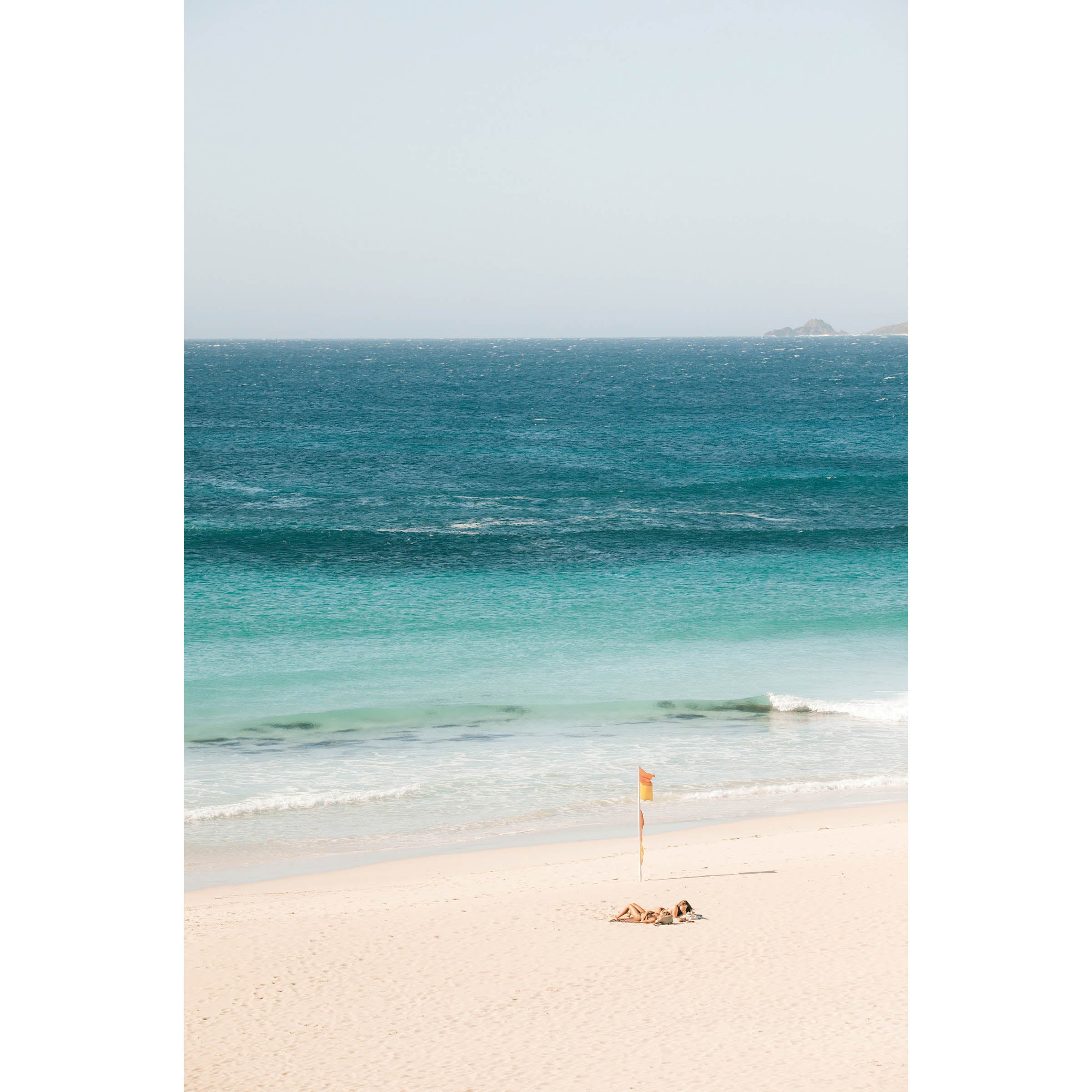 Colour photo of two girls sunbathing on Smiths Beach in Western Australia, next to the lifeguard red and yellow flag as the ocean gently rolls in. Available framed in black, white and oak.