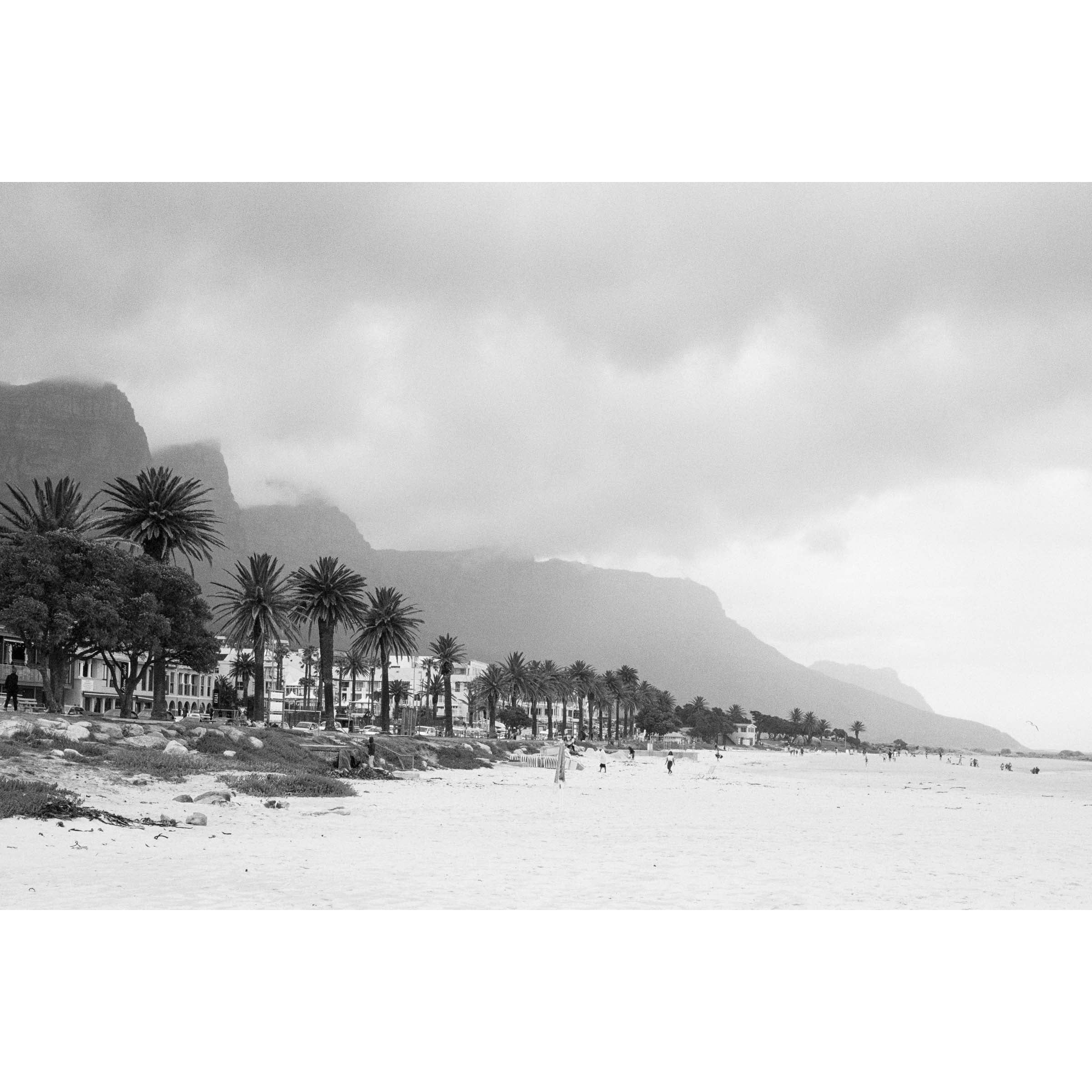 Horizontal black and white photo of Camps Bay Beach in Cape Town, South Africa. The palms line the edge of the sand and the mountains rise in the background behind clouds. Shot on film it has a moody, nostalgic feel. Fine art photography prints, available framed in black, white or oak.