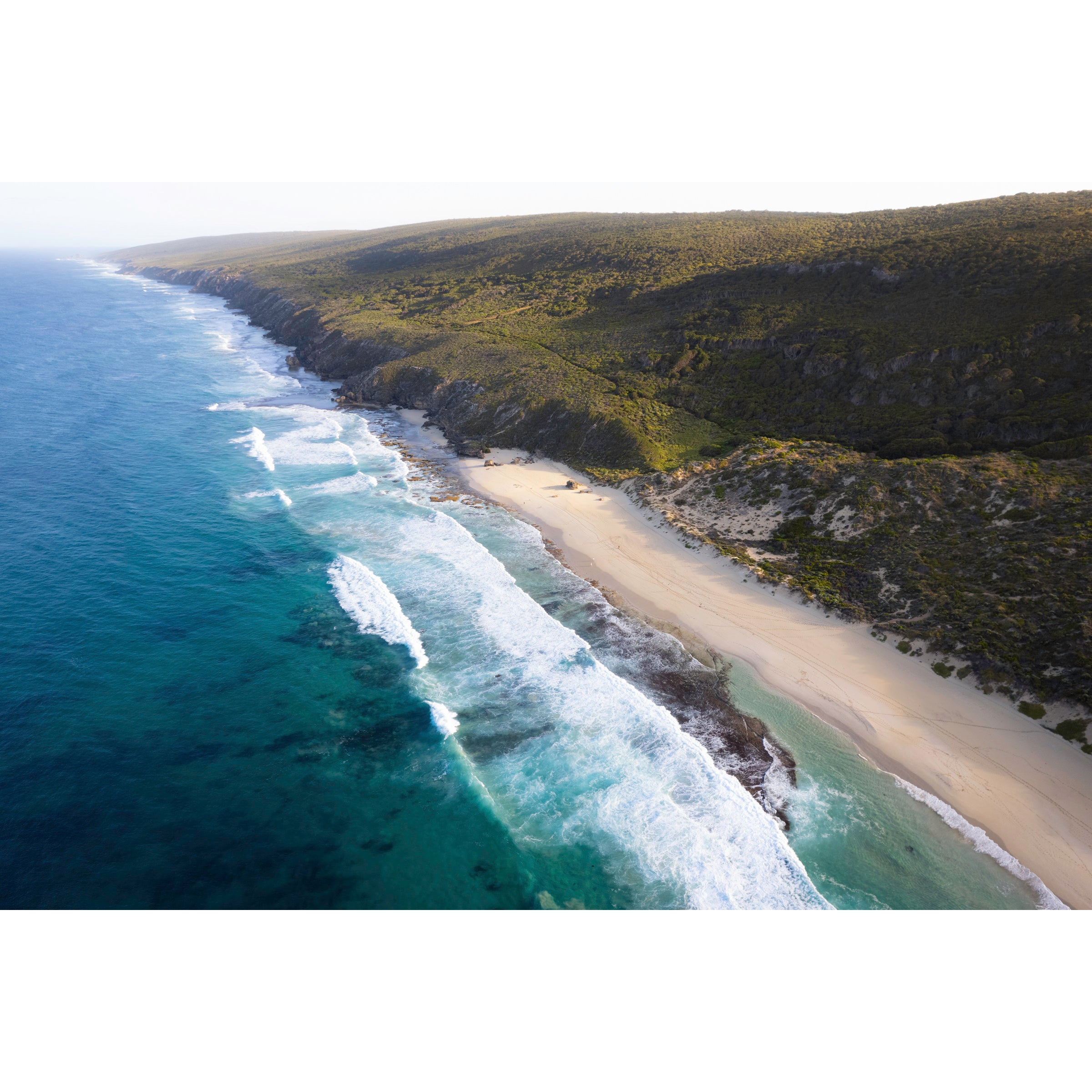 Horizontal drone photo of the far end of Yallingup Beach with the waves rolling in to the white sand and the bushland stretching into the distance. Original fine art photography of Western Australia, available as prints and framed wall art. Perfect for a coastal home or beach house.