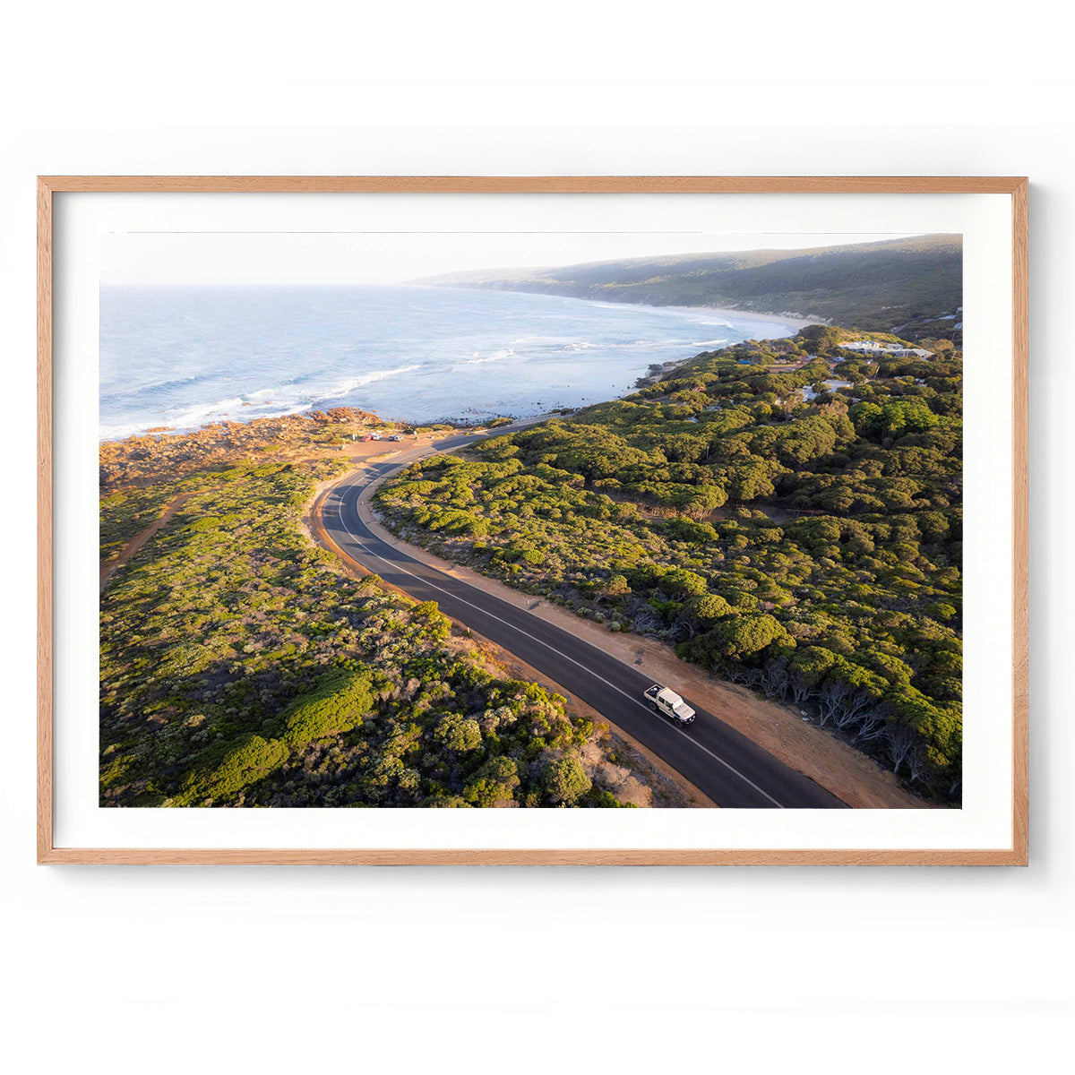 Horizontal photo of the road down to Yallingup beach in Western Australia. Taken from above at sunrise so it has a golden glow, with a car driving up the hill and the ocean in the background. Perfect for the coastal home or beach house. Available as fine art photography prints and framed wall art.