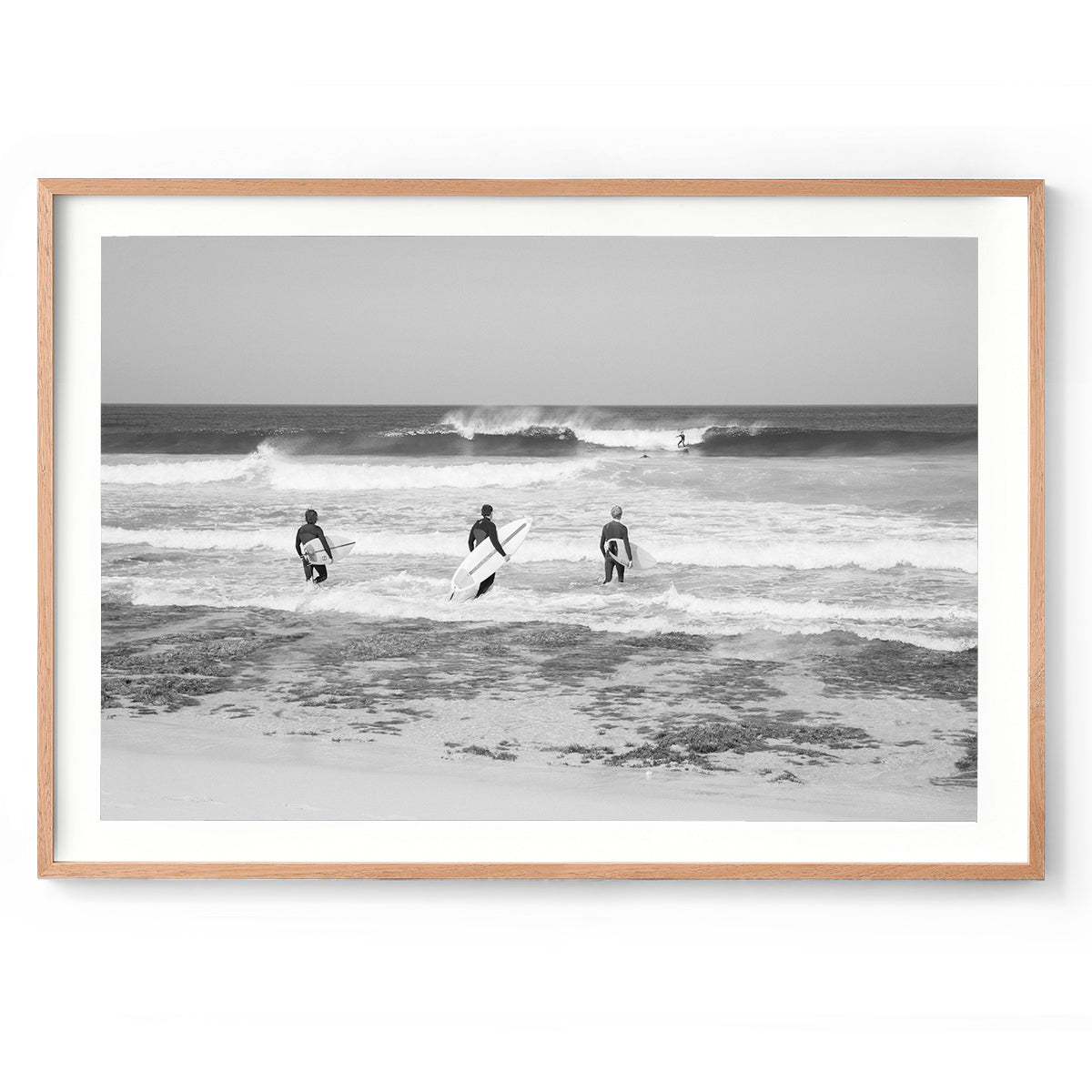 Horizontal black and white photo of three surfers walking into the ocean at Rabbits Break, Yallingup Beach, Western Australia. They have their surfboards under their arms and a wave rolls in with a surfer on it. Fine art photography prints, available framed in black, white or oak.