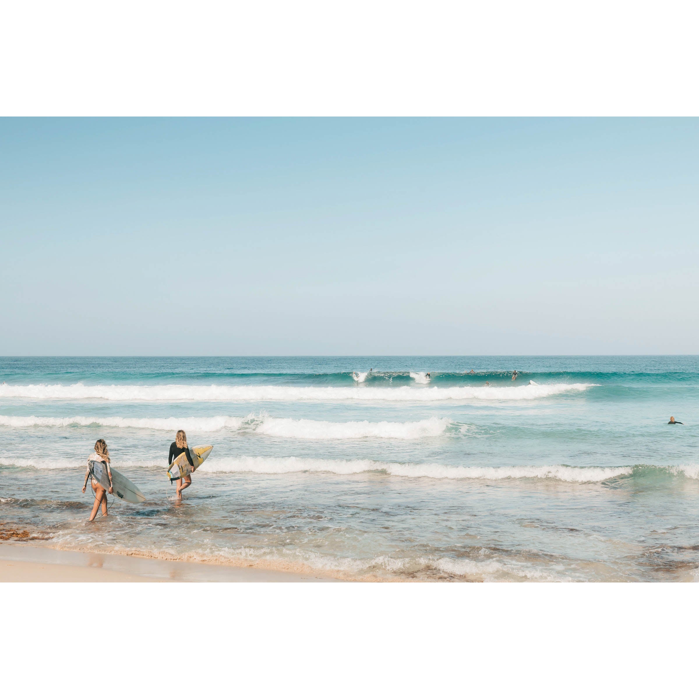 Framed horizontal photo of two female surfers walking into the ocean at Yallingup Beach in Western Australia holding their surfboards under their arms. Waves are rolling in with surfers dotted throughout the soft blue water. Available framed in oak, black or white.