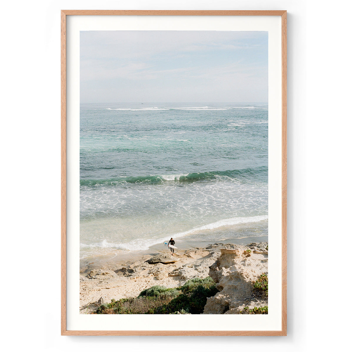 Photo of a surfer coming out of the ocean at Surfer's Point in Margaret River, Western Australia. Shot on film with soft colours, there are rocks in the foreground and the ocean stretches out towards the horizon with waves rolling in. Available framed in black, white or oak.