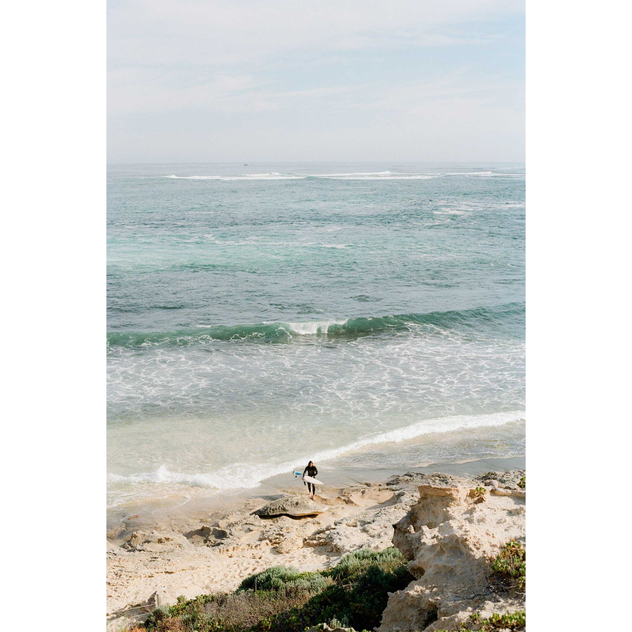 Photo of a surfer coming out of the ocean at Surfer's Point in Margaret River, Western Australia. Shot on film with soft colours, there are rocks in the foreground and the ocean stretches out towards the horizon with waves rolling in. Available framed in black, white or oak.
