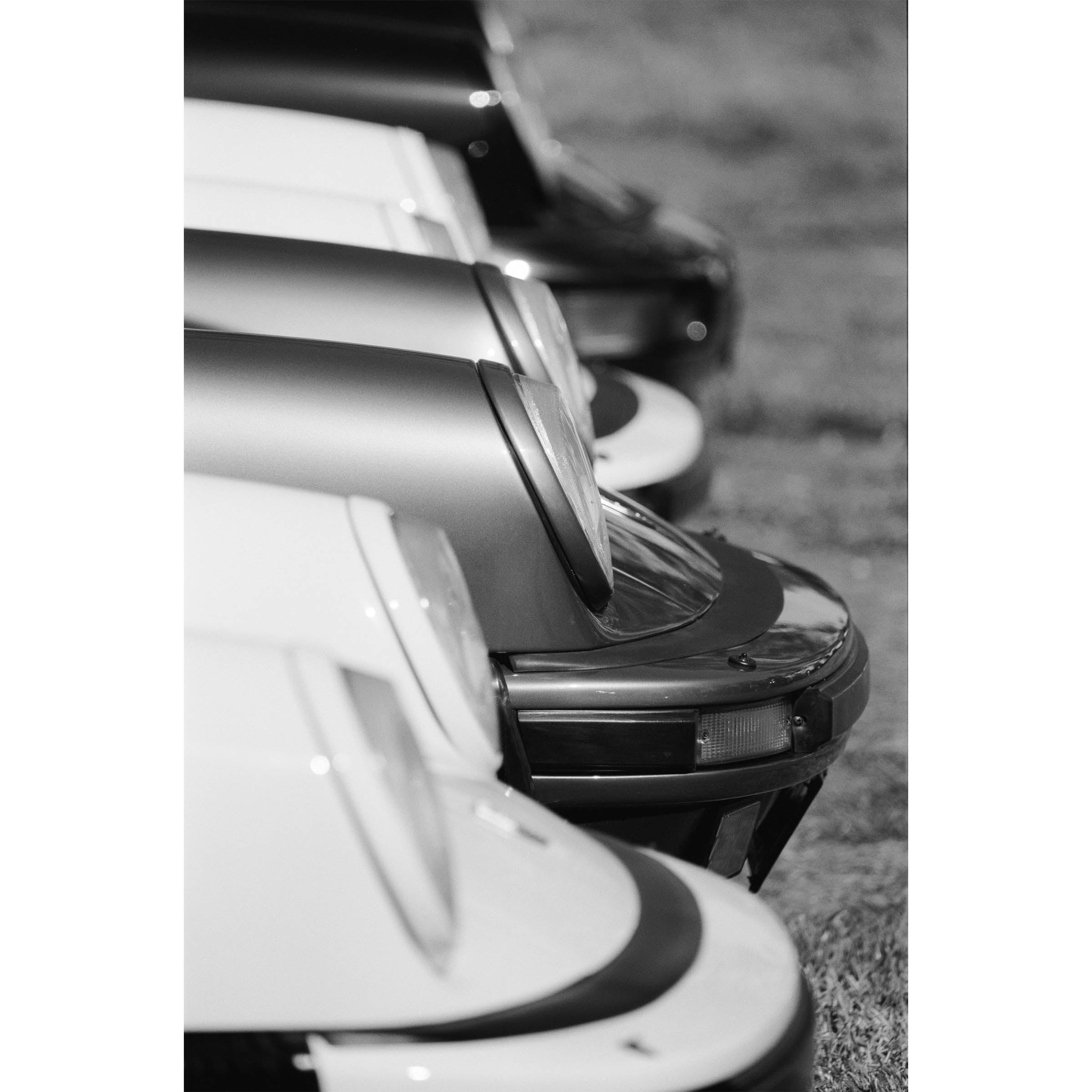 Vertical black and white photo of the front of multiple Porsche 911s from the side all lined up showing just the iconic round headlights and bumpers. Fine art photography prints, available framed in black, white and oak.