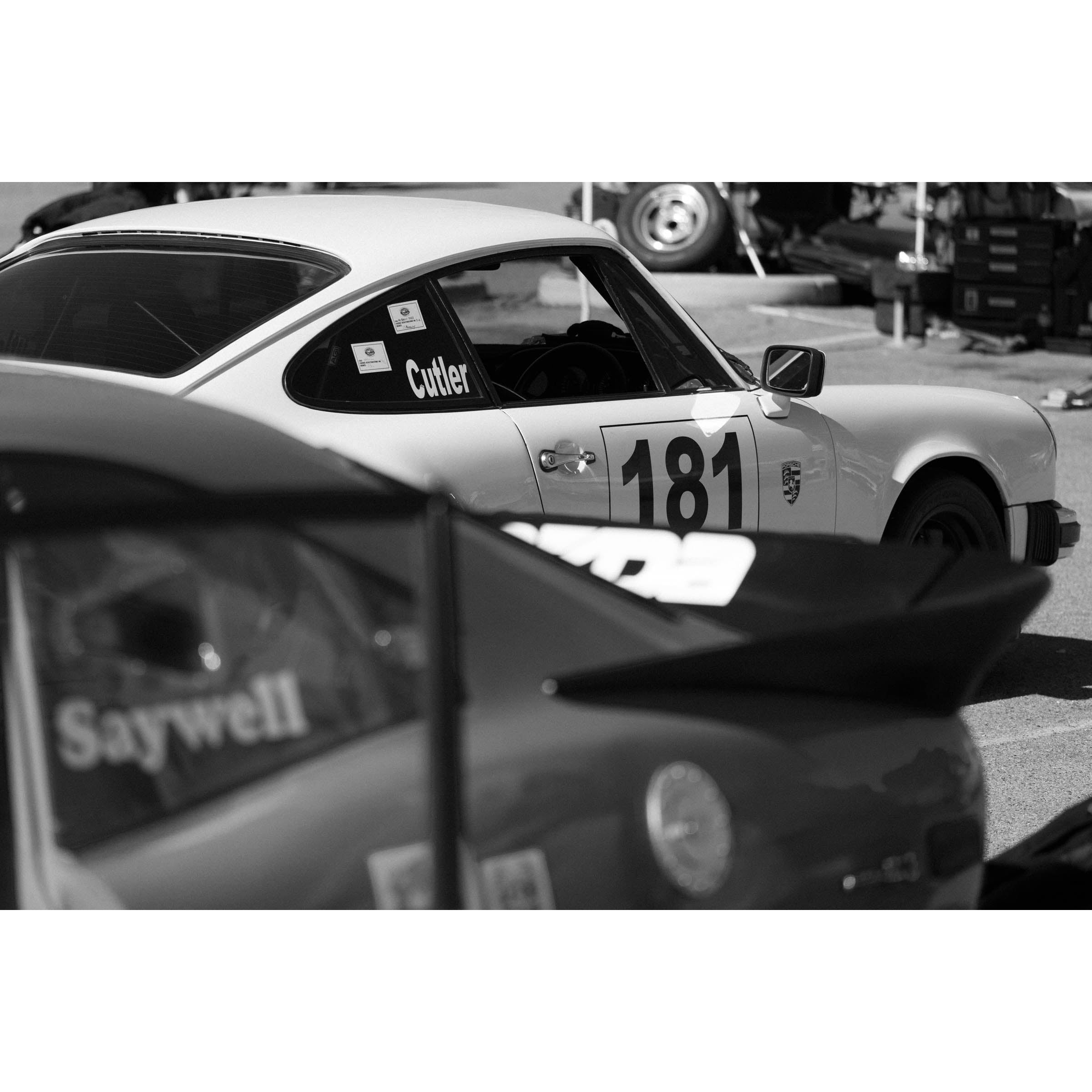 Horizontal black and white photo of a Porsche 911 with a race number on the side waiting in the pits to race again. Fine art photography prints, available framed in black, white or oak.