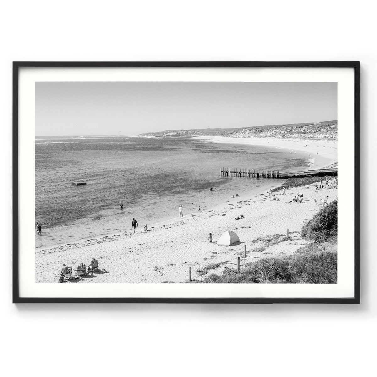 Black and white photo of Gnarabup Beach in Margaret River, Western Australia. It's a sunny day and there are people on the sand and in the water, a tent on the beach and the jetty sits out in front of the beach curving around under the cliffs. Available framed in black, white or oak.