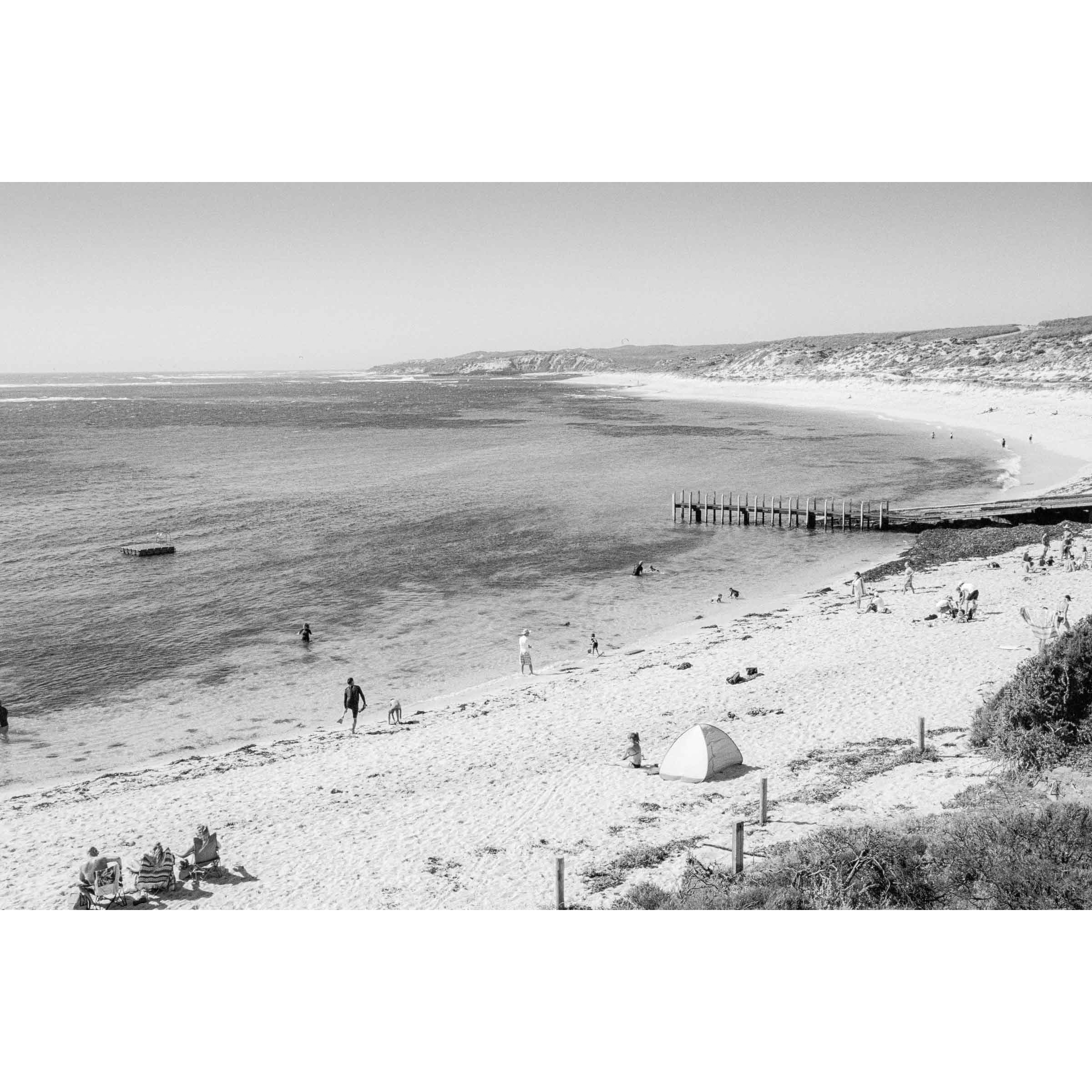 Black and white photo of Gnarabup Beach in Margaret River, Western Australia. It's a sunny day and there are people on the sand and in the water, a tent on the beach and the jetty sits out in front of the beach curving around under the cliffs. Available framed in black, white or oak.