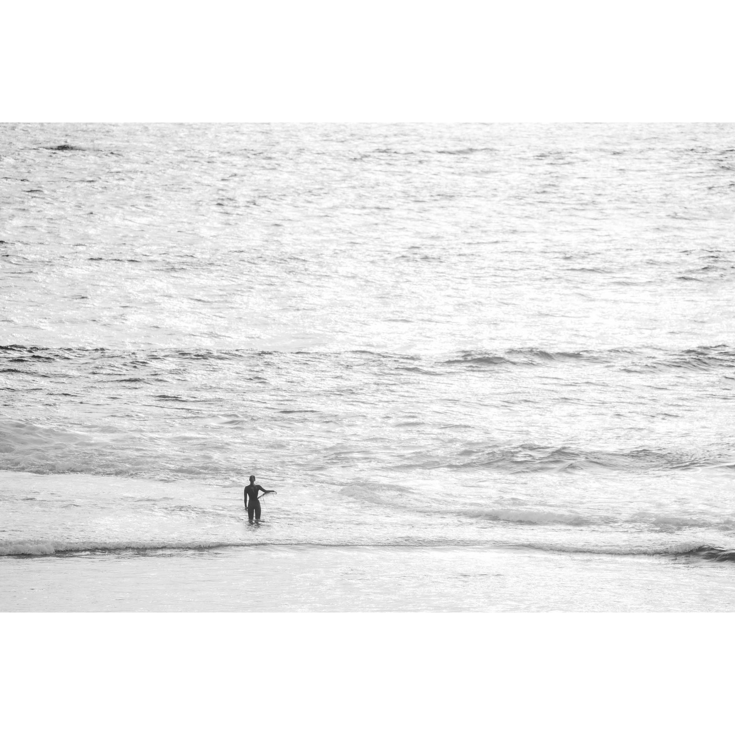 Black and white photo of a female surfer holding a surf board on her hip looking out whilst standing in the ocean at Yallingup Beach. Fine art photography prints, available framed in black, white or oak.