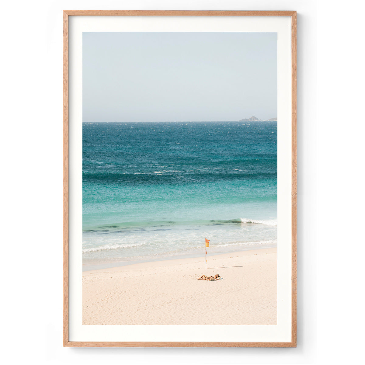 Colour photo of two girls sunbathing on Smiths Beach in Western Australia, next to the lifeguard red and yellow flag as the ocean gently rolls in. Available framed in black, white and oak. 
