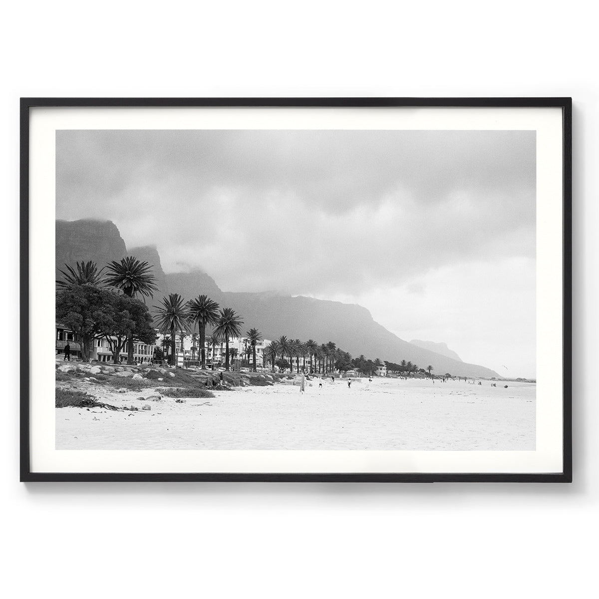 Horizontal black and white photo of Camps Bay Beach in Cape Town, South Africa. The palms line the edge of the sand and the mountains rise in the background behind clouds. Shot on film it has a moody, nostalgic feel. Fine art photography prints, available framed in black, white or oak.