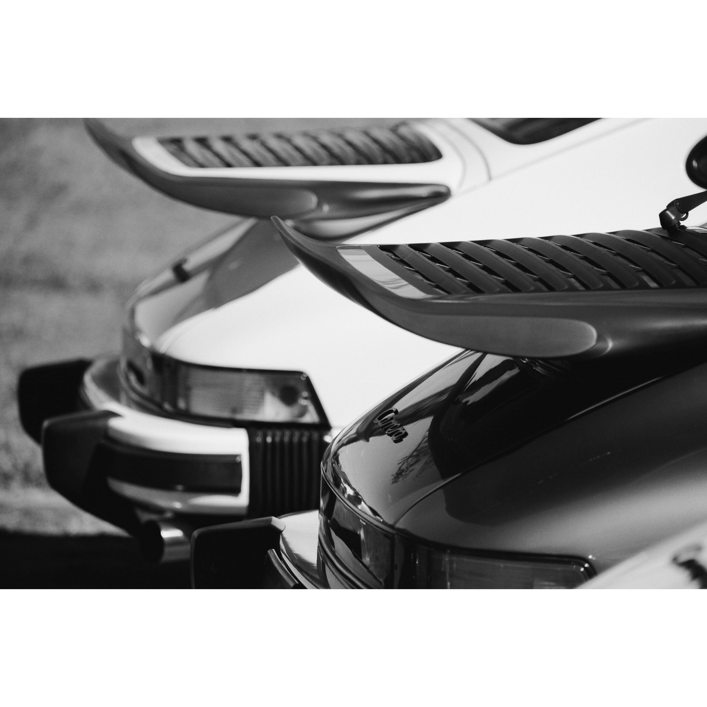 Black and white photo of the back of two Porsche 911s showing the iconic whale tail spoilers. Fine art photography prints, available framed in black, white or oak.